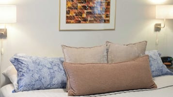 A neatly made bed with patterned pillows, a framed abstract painting on the wall, and wall-mounted lamps.