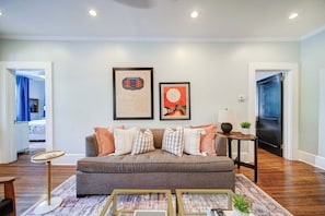 A living room with a grey sofa, two framed artworks, a small side table with a lamp, and a wooden floor.
