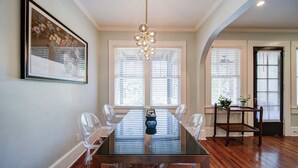 A dining area with a glass table, clear chairs, a chandelier, and large windows with blinds. - The Home Collection: Historic Myers Park 2BR/2BA | Step-Free | Screened Porch (Charlotte)