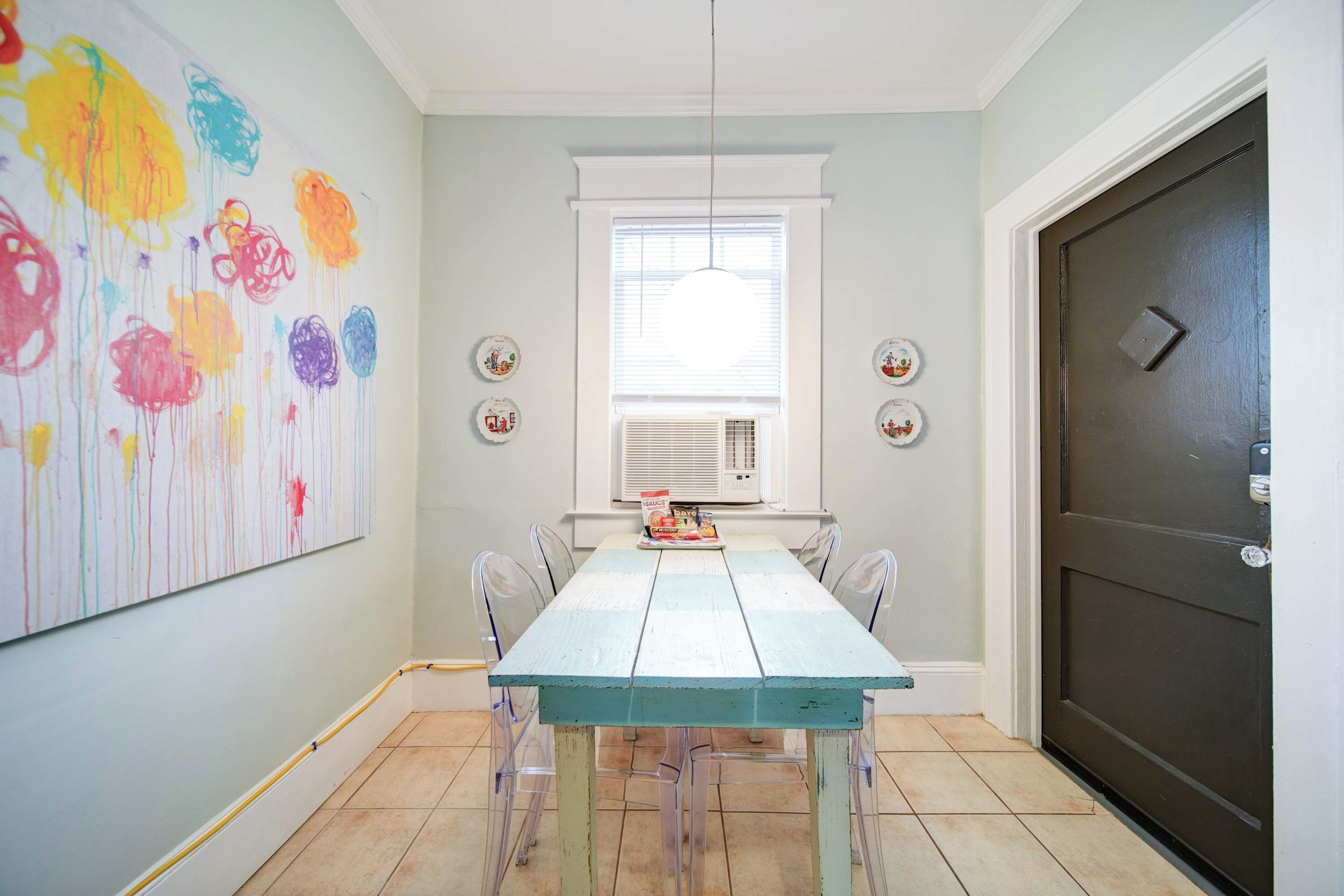 A dining area with a light blue table and chairs, a large abstract painting on the wall, and a dark door to the right.