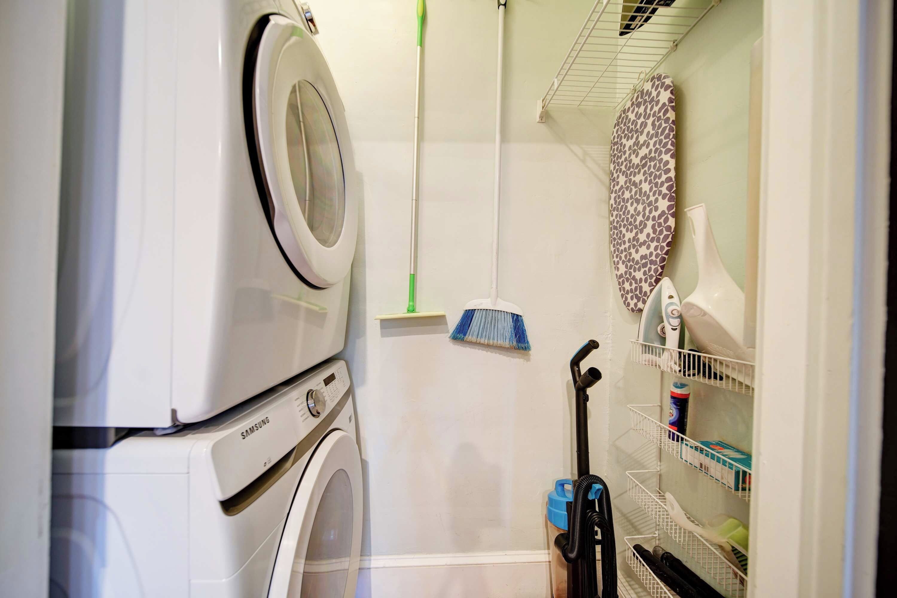 Een kleine wasruimte met een wasmachine, een droogrek, een bezem en een plank met schoonmaakspullen.