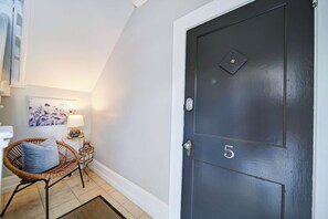 Une porte sombre portant le numéro 5, une chaise en osier avec un coussin, une petite table avec une lampe et un tableau accroché au mur.