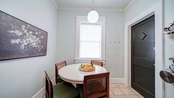 Uma área de refeições com uma mesa redonda, duas cadeiras, uma cesta de frutas, uma luminária no teto e uma porta.