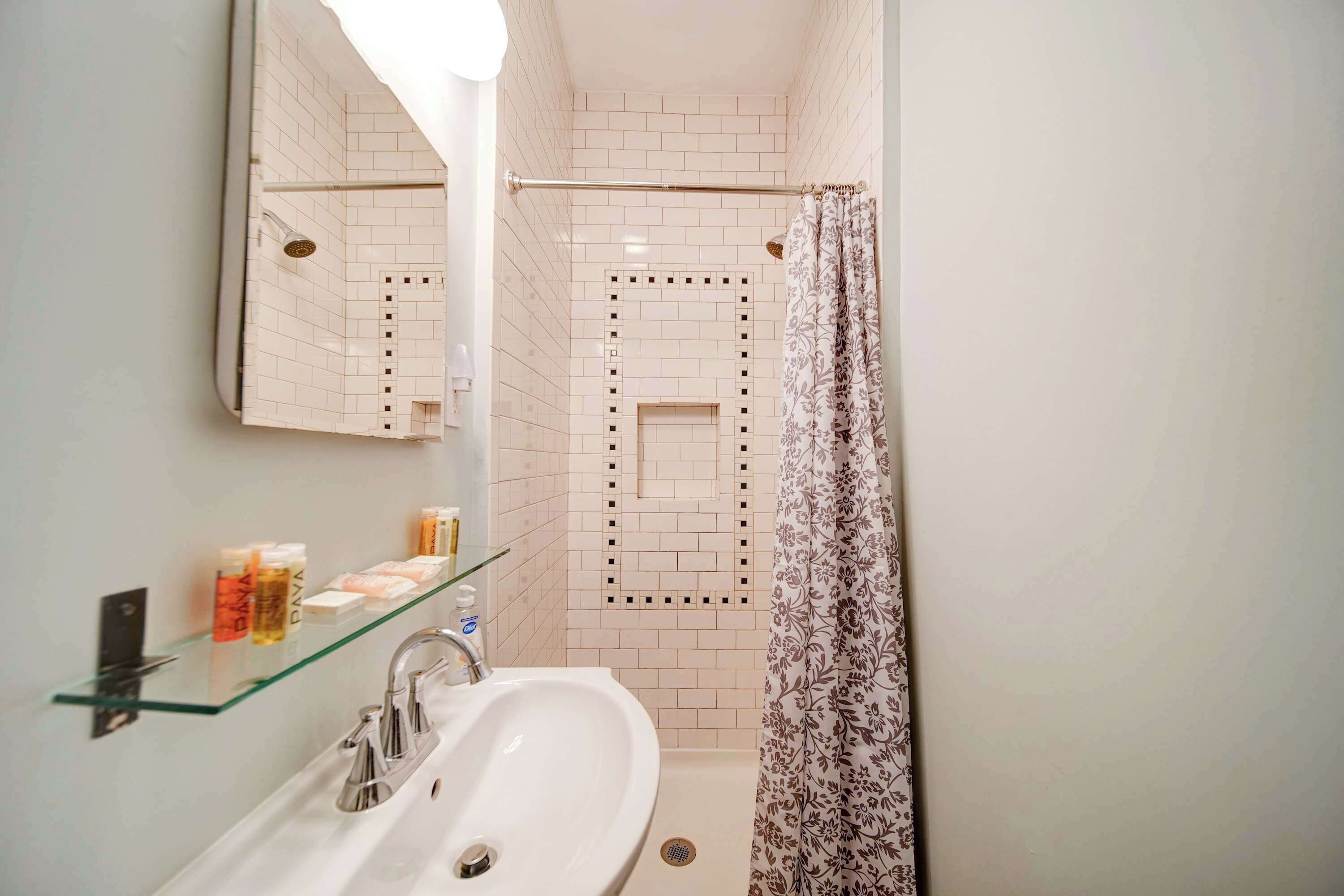 Un baño con repisa de vidrio que contiene artículos de tocador, un lavamanos blanco, un espejo y una ducha con cortina estampada.