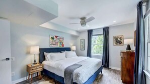 A modern bedroom with a large bed, a ceiling fan, a nightstand with a lamp, and a view of greenery outside.