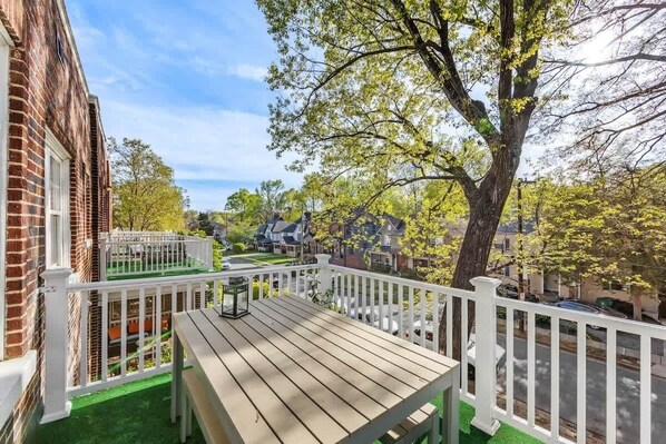 A wooden deck with a table and chairs, surrounded by a white fence, overlooking a residential neighborhood with houses and trees.
