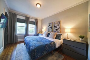 Un dormitorio con una cama grande, dos mesitas de noche, un buró, un televisor y una ventana con cortinas.