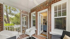 Ein überdachter Balkon mit Ziegelwänden, Fenstern mit weißen Rahmen und einer Glastür, die zu einem Raum führt. Wicker-Möbel und ein Glastisch sind ebenfalls vorhanden.