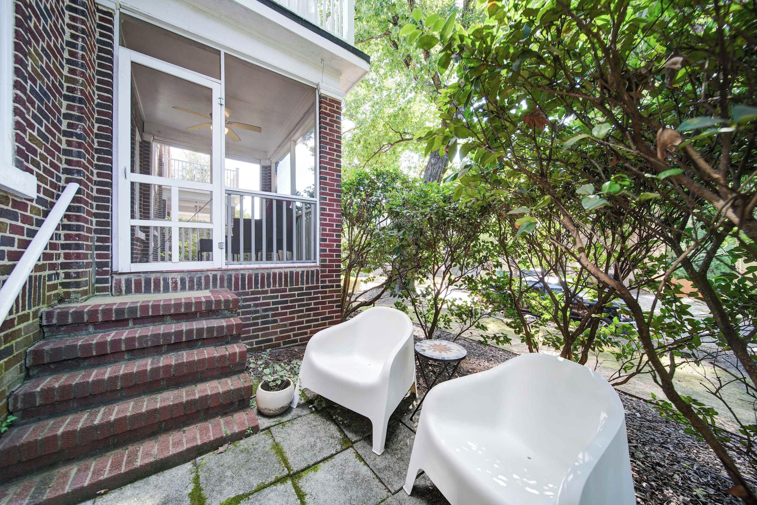 A brick house with a porch, steps leading to a door, and a small garden area with a white chair and a potted plant.
