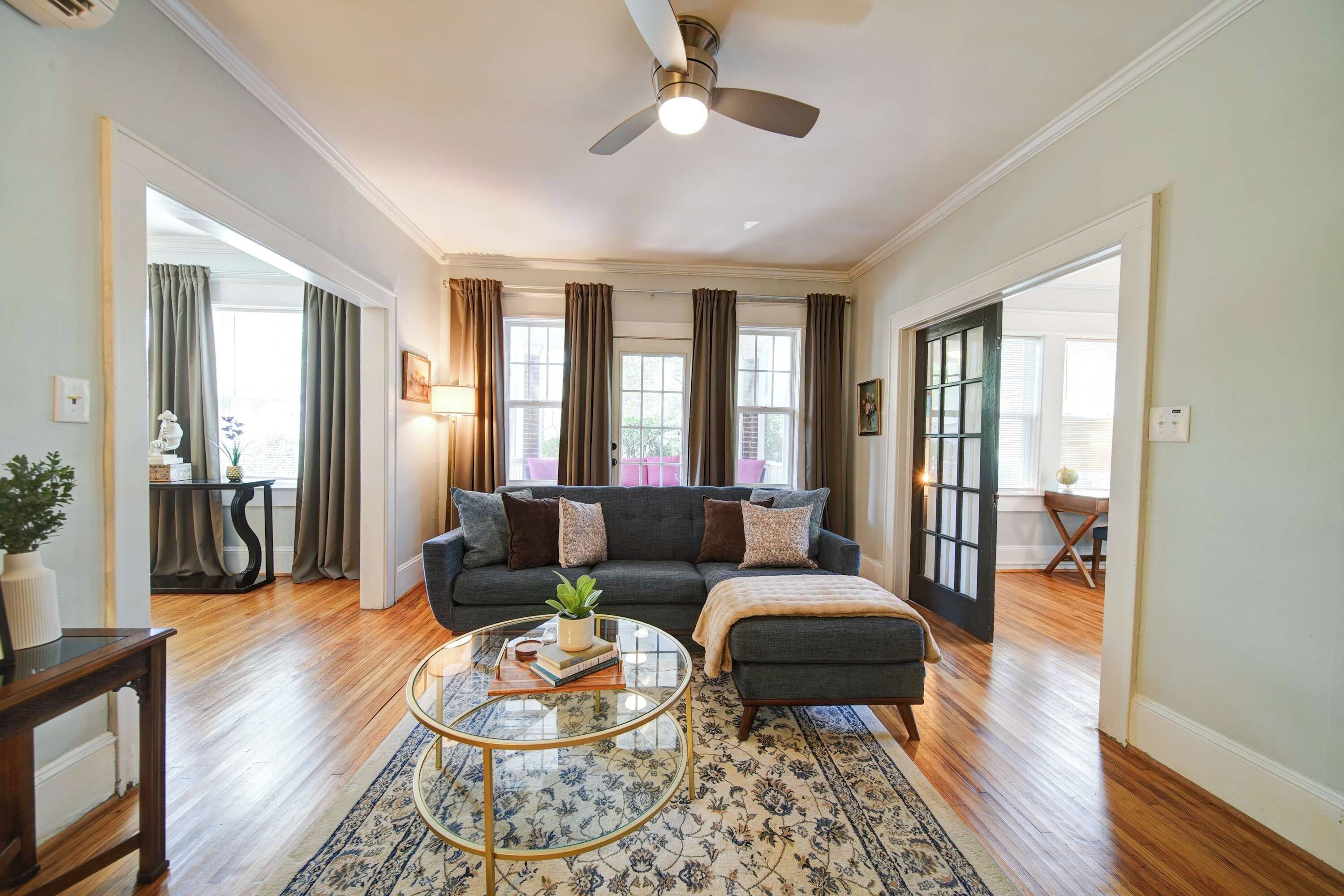A spacious living room with a dark grey sofa, a round glass coffee table, and a ceiling fan.