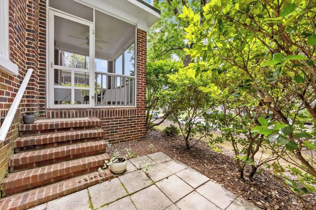 A brick house with a porch, steps leading to a door, and a small garden area with a tree and potted plants.