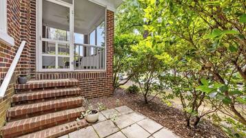 A brick house with a porch, steps leading to a door, and a small garden area with a tree and potted plants.