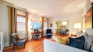 A living room with a sofa, a chair, a TV, and a dining area with a table and chairs. - The Home Collection: Hawthorne 1BR King Suite | Private Balcony | Walk to Novant (Charlotte)