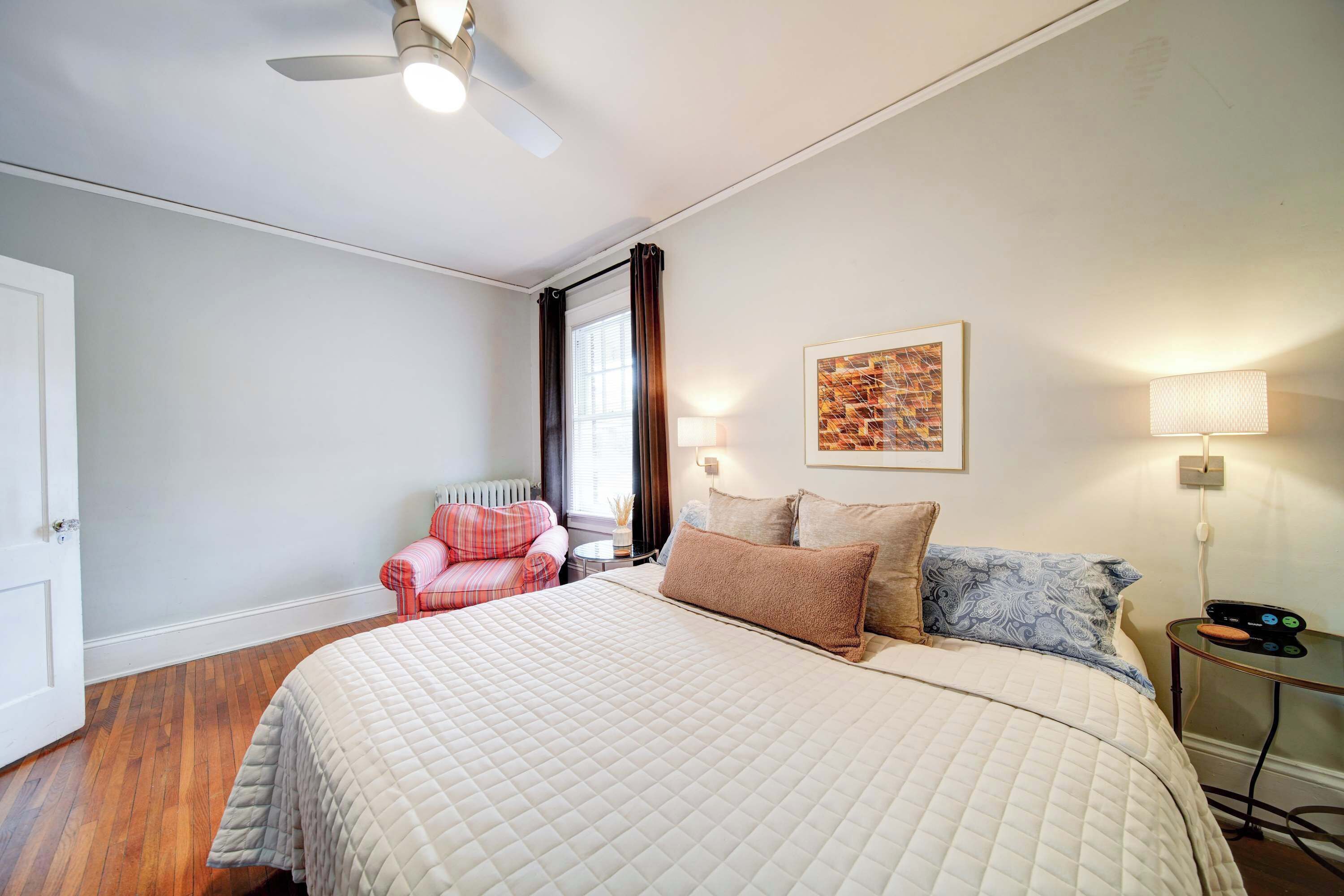 A bedroom with a large bed, a ceiling fan, a window with curtains, a bedside table with a lamp, and a wall-mounted artwork.