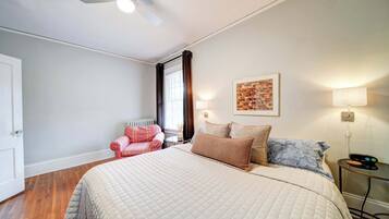 A bedroom with a large bed, a ceiling fan, a window with curtains, a bedside table with a lamp, and a wall-mounted artwork.