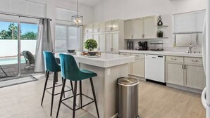 A modern kitchen with white cabinets, a central island with bar stools, a coffee machine, and a view of the outdoors. - Stunning 5BD Escape w/ Billiard Room & Pool! (North Las Vegas)
