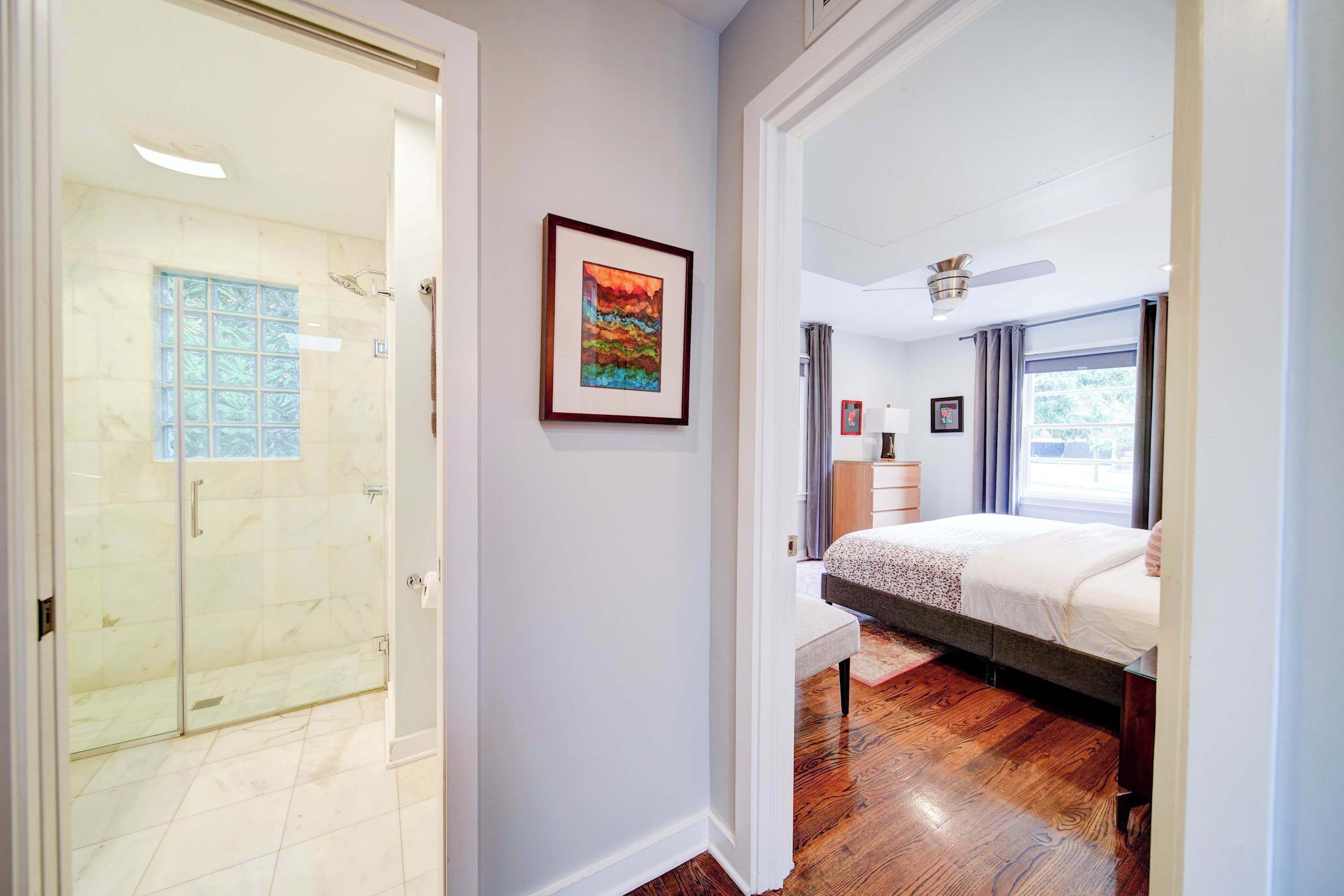 A hotel room with a glass shower, a bed with a headboard, a bedside table, and a window with curtains.