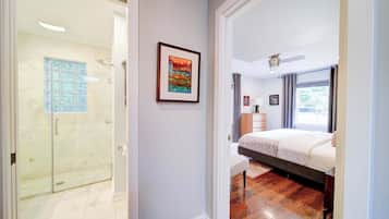 A hotel room with a glass shower, a bed with a headboard, a bedside table, and a window with curtains.