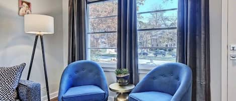 A room with two blue armchairs, a floor lamp, a small table with a plant, and a view of a tree-lined street through large windows.