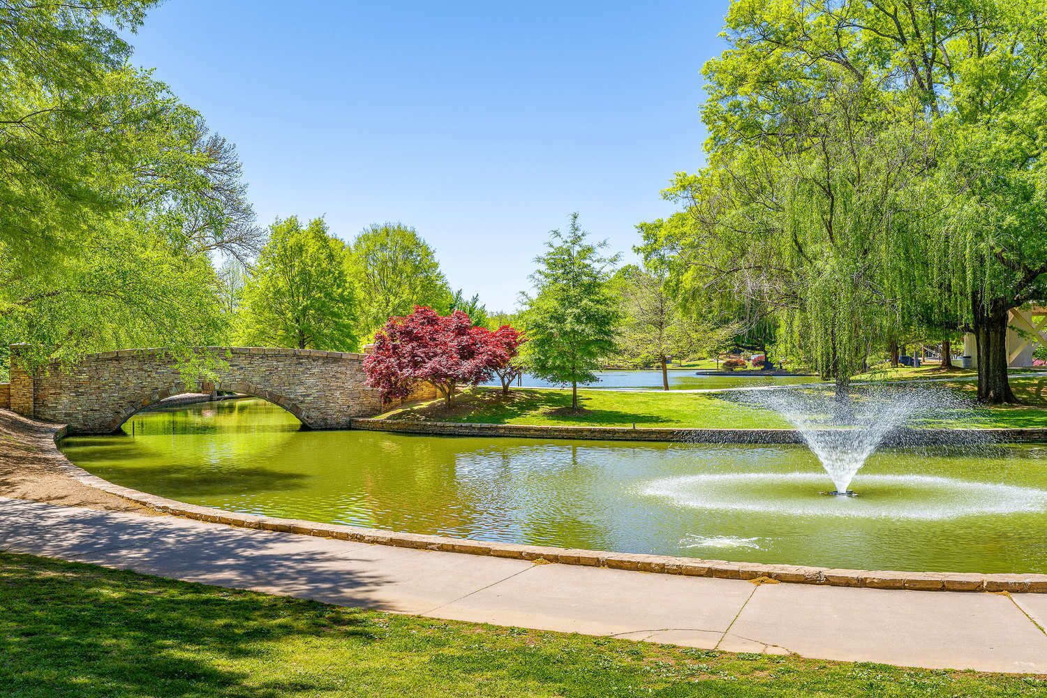 A park with a stone bridge, a pond with a fountain, and trees.