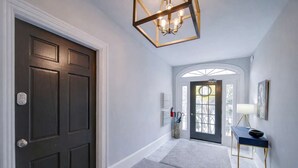 A hallway with a dark door, a light-colored wall, a geometric ceiling light fixture, a blue console table with a lamp, and a door with a glass panel.