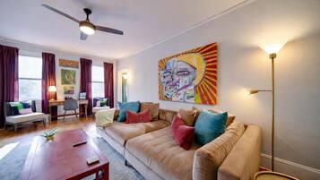 A living room with a beige sofa, a wooden coffee table, a desk with a chair, and a ceiling fan.