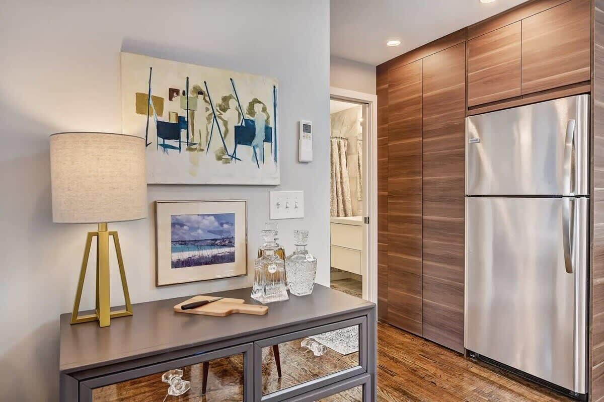 A modern kitchen with a stainless steel refrigerator, wooden cabinets, a framed painting, a lamp, and a console table with glassware.