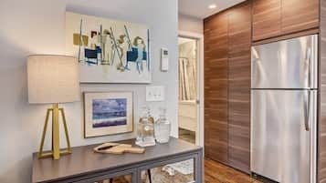 A modern kitchen with a stainless steel refrigerator, wooden cabinets, a framed painting, a lamp, and a console table with glassware.