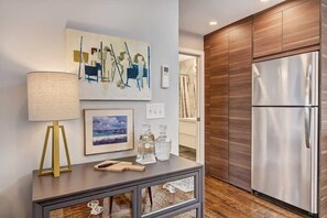 A modern kitchen with a stainless steel refrigerator, wooden cabinets, a framed painting, a lamp, and a console table with glassware.