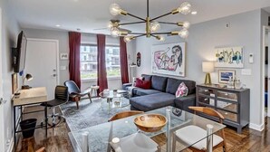 Een moderne woonkamer met een grijze bank, een glazen salontafel en een houten eettafel. Er hangt een kroonluchter met meerdere lampen, een televisie is aan de muur bevestigd en er is een raam met gordijnen.