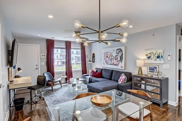 A modern living room with a grey sofa, a glass coffee table, and a wooden dining table. There is a chandelier with multiple bulbs, a TV mounted on the wall, and a window with curtains.