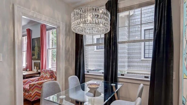 A dining area with a glass table and chairs, a chandelier, and large windows with curtains.