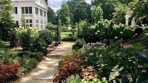 A garden pathway leading to a white building with columns, surrounded by lush greenery and flowering plants. - The Home Collection: Historic Myers Park 2BR · Near Atrium Health (Charlotte)