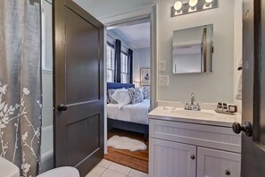 Un baño con lavamanos blanco, espejo y una puerta que da a un dormitorio con cama y ventana.