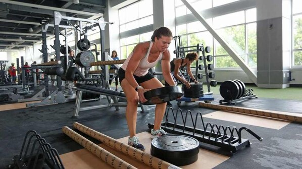 Uma academia com equipamentos de musculação, incluindo barras e pesos, e uma pessoa executando levantamento terra.