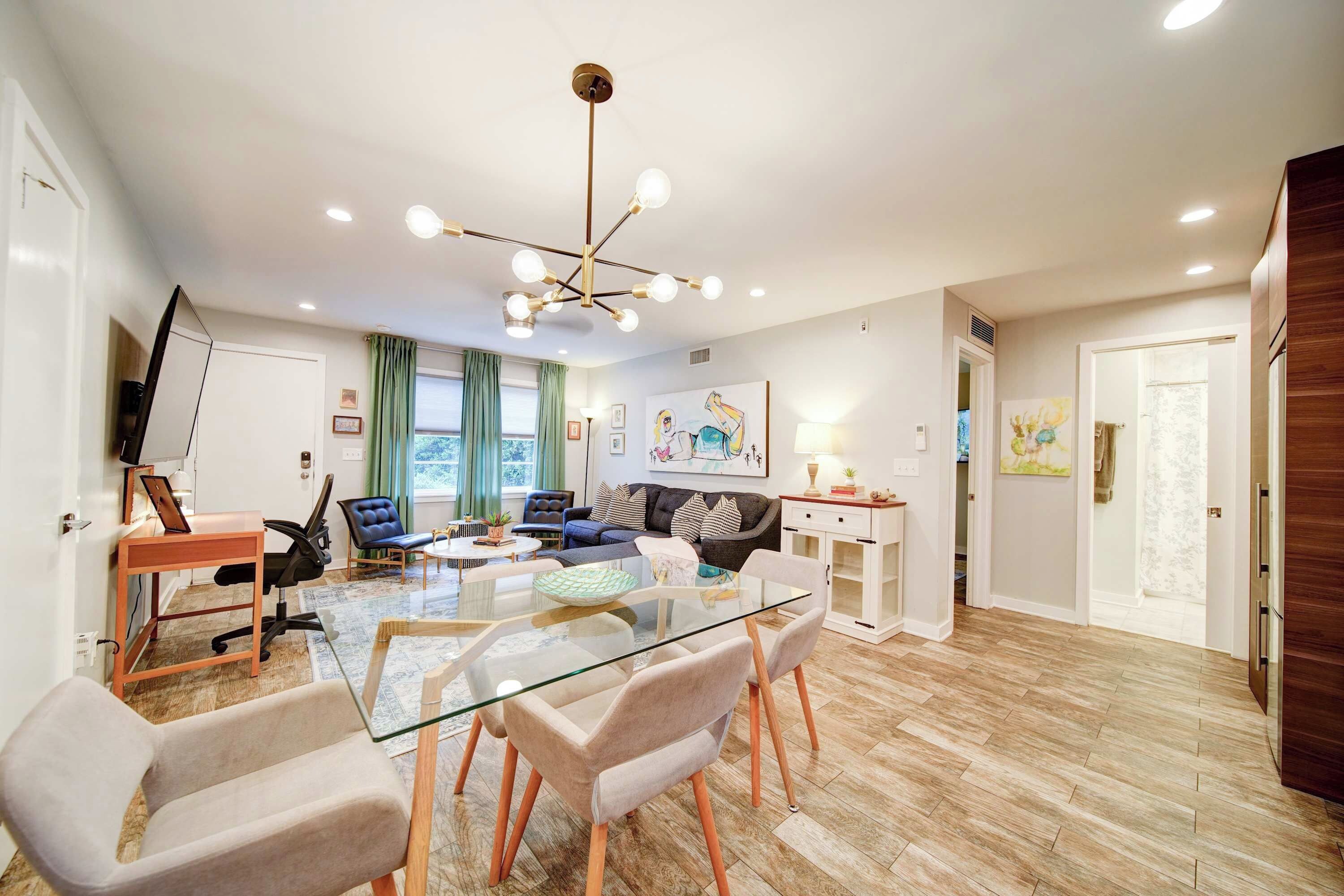 A modern living room with a glass-top dining table, leather chairs, a sofa, and a ceiling light fixture.