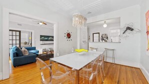 A modern living room with a white dining table, clear chairs, a blue sofa, and a chandelier.