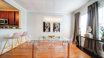 A modern dining area with a glass table and clear chairs, a kitchen with wooden cabinets, and a decorative chandelier.
