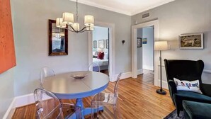 A dining area with a round table, clear chairs, and a chandelier. There is a framed picture on the wall and a lamp on a side table.