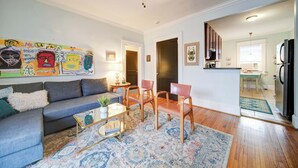 Un salon avec un canapé, des fauteuils, une table basse et un tapis. Au fond, on aperçoit une cuisine équipée d’un réfrigérateur et d’un four à micro-ondes.