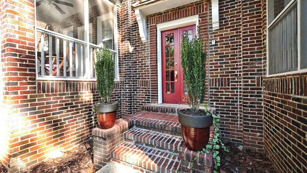 Un bâtiment en briques avec une porte rouge, deux plantes en pot sur les marches et une fenêtre grillagée.