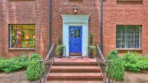 Un bâtiment en briques avec une porte bleue, des marches menant à l’entrée et un petit jardin agrémenté de plantes en pot.