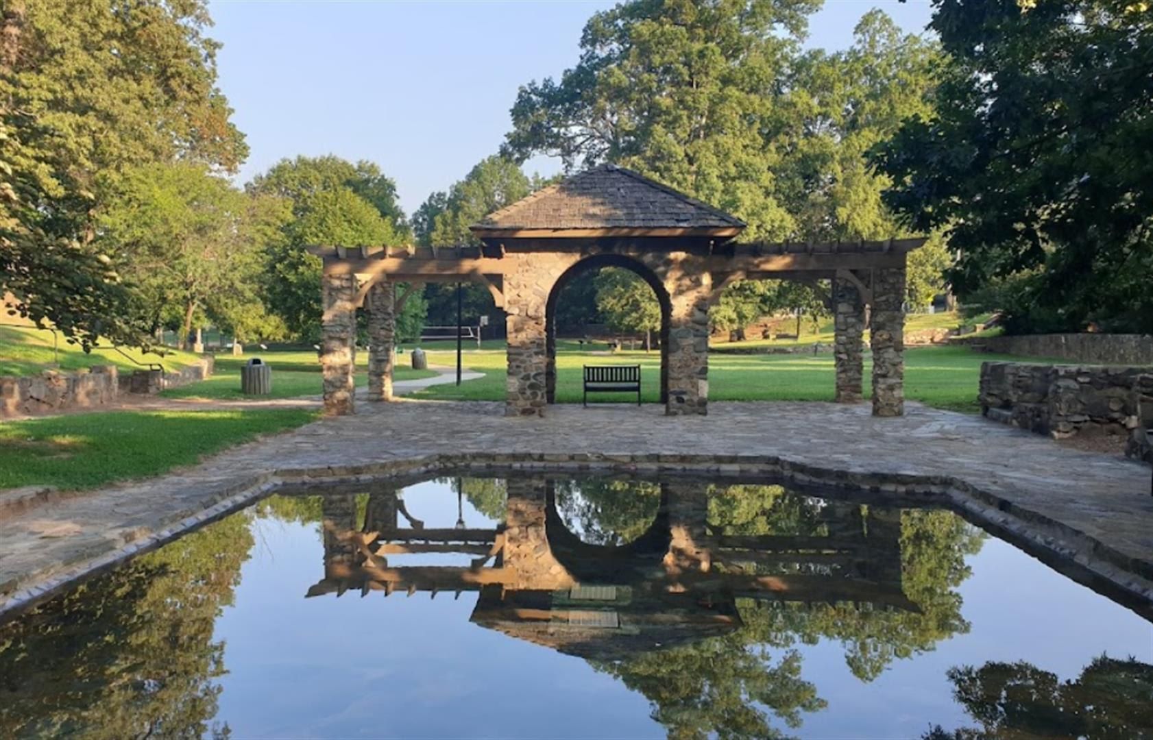 A stone gazebo with a tiled roof, surrounded by a reflective water feature and greenery.