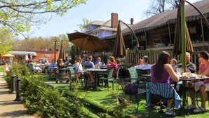 Un área de comedor al aire libre con mesas y sillas, rodeada de vegetación y un edificio al fondo.