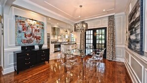 A spacious dining area with a glass table and chairs, a black console with drawers, a large abstract painting on the wall, and a chandelier.