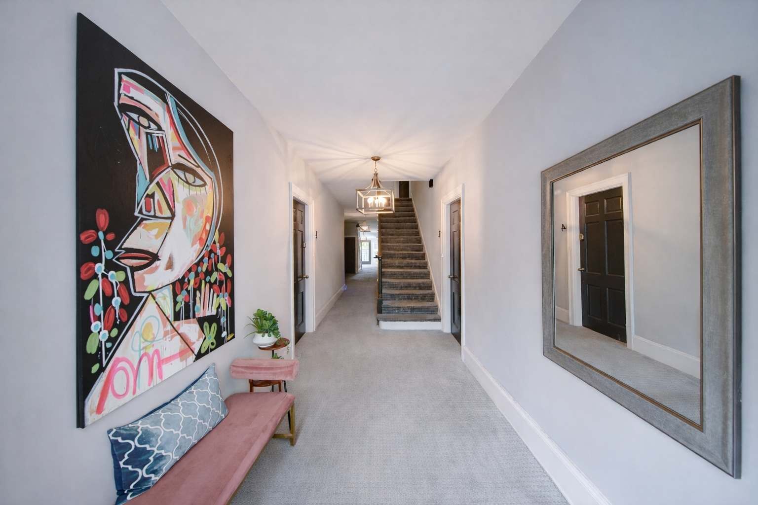 Un couloir avec un escalier, un banc, une plante en pot et un grand tableau abstrait accroché au mur.