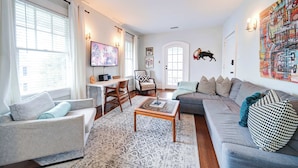 A living room with a grey sofa, a wooden coffee table, and a patterned rug. There is a television on a stand, a chair, and a door with a bull mural. - Modern 2BR Elizabeth Suites | Parking & Private Balcony | near Uptown (Charlotte)