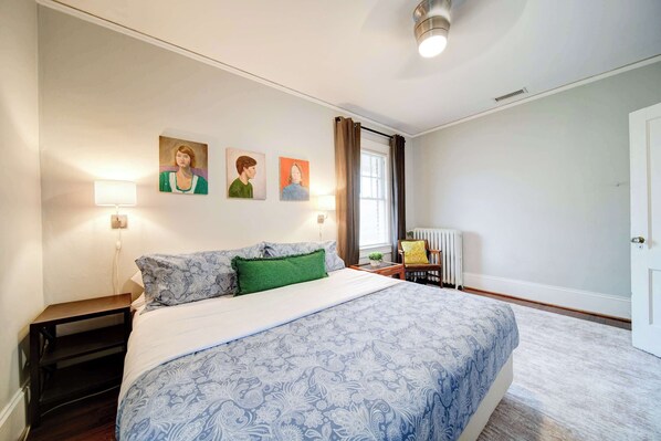 A bedroom with a large bed, a nightstand, a lamp, a chair, and three paintings on the wall.