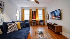 Un soggiorno con un divano blu scuro, un tavolino in vetro, una ventola a soffitto, una TV e un quadro appeso al muro.
