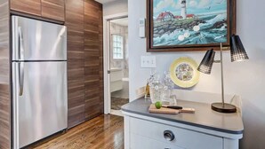 A modern kitchen with a stainless steel refrigerator, wooden cabinets, a painting of a lighthouse, and a small dining area with a table and chairs.